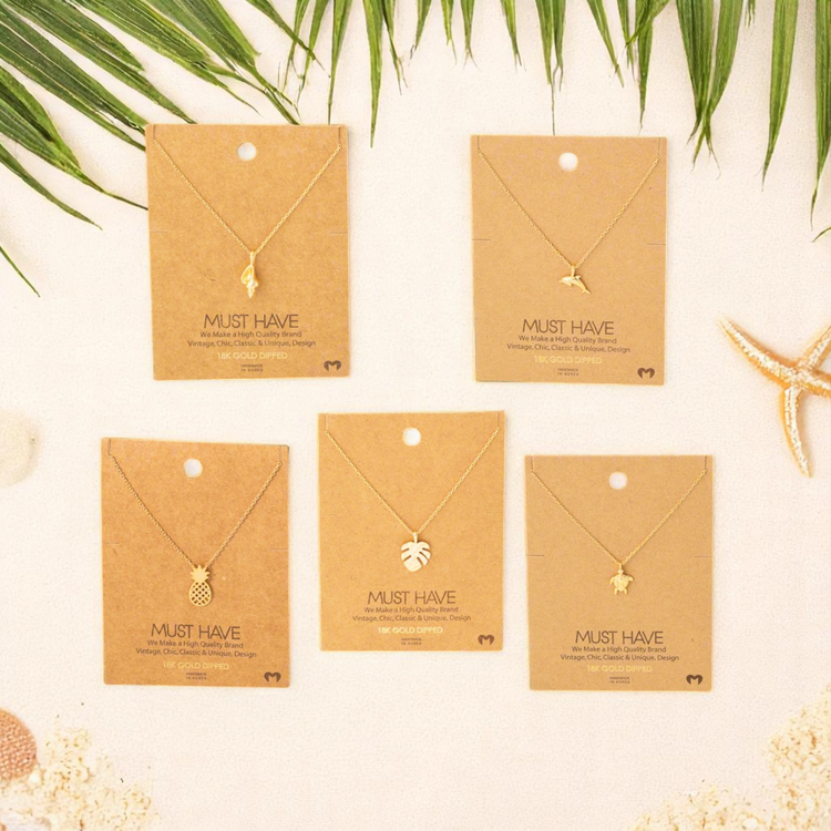 Flat lay of gold-dipped beach-themed necklaces on kraft display cards with a luxury coastal background featuring palm leaves and soft sand tones