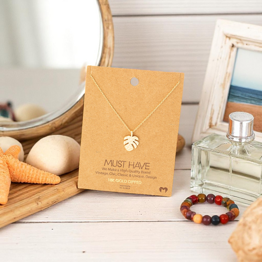 Monstera leaf pendant necklace displayed on a jewelry card in front of a vanity mirror with beach-inspired decor and soft natural lighting.
