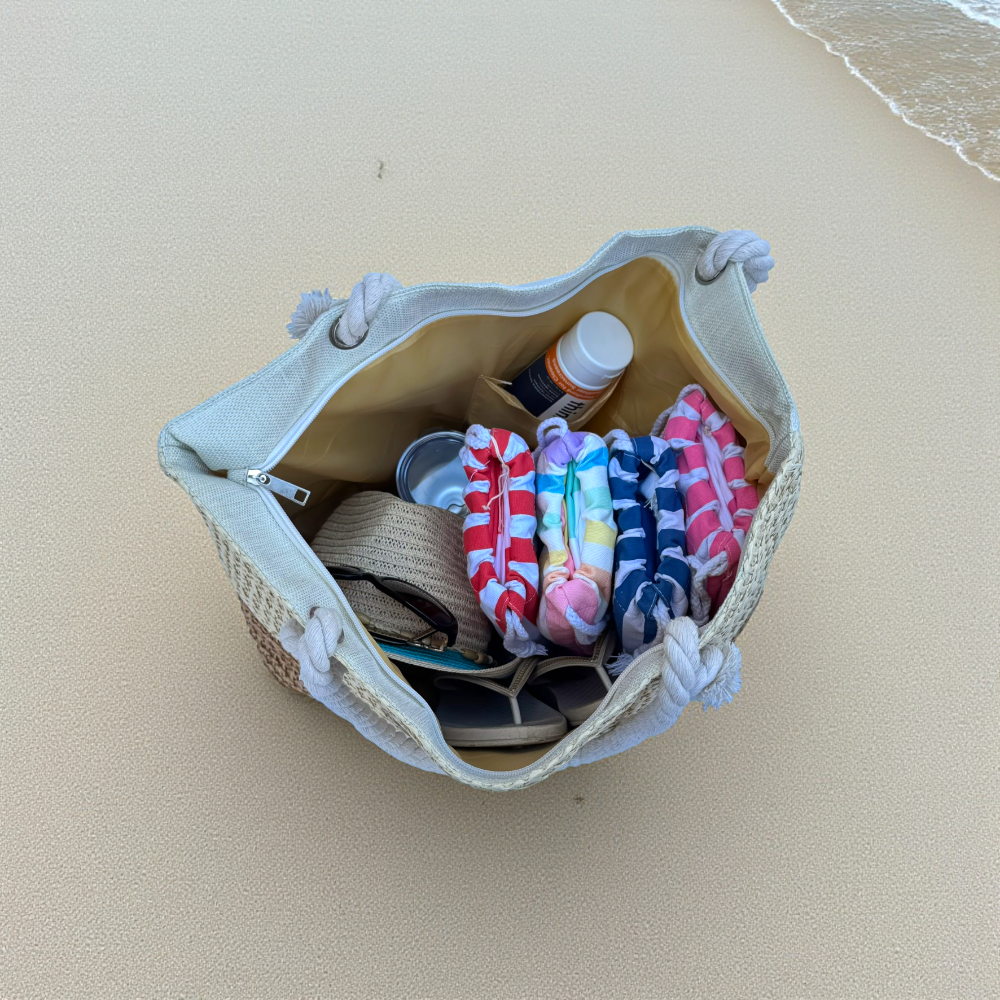 Top-down view of straw beach tote bag fully packed with beach essentials on pale sand with ocean wave visible on the edge