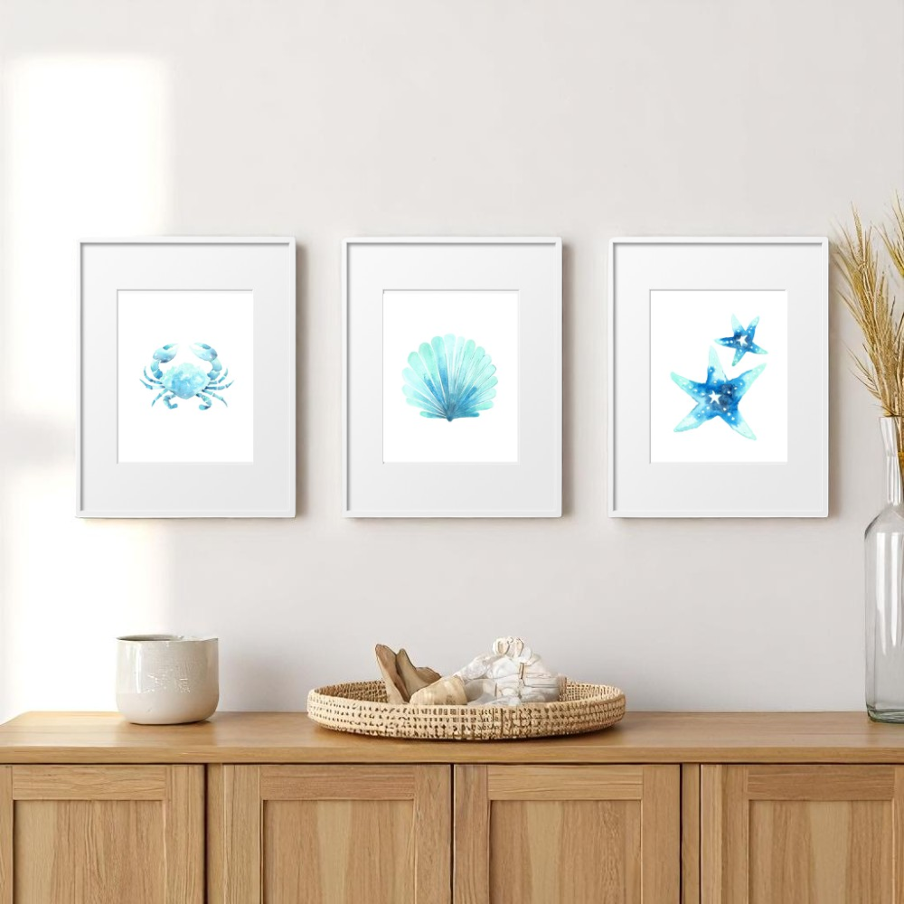 Three framed 8x10 beach prints on a light wall above a natural wood console table, decorated with a woven tray, beach shells, neutral ceramic, and dried coastal grass.