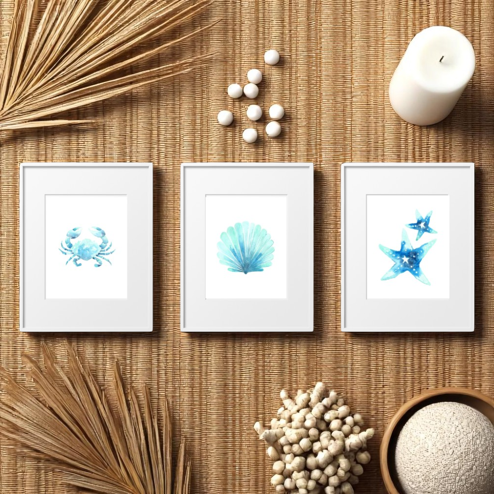 Flat lay of three framed beach art prints with crab, shell, and starfish designs on a woven rattan surface, styled with dried palm leaves, candle, and natural coastal decor.