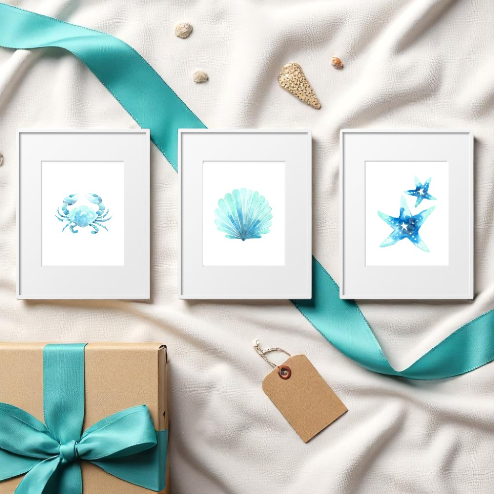 Flat lay of three framed beach prints styled on soft white fabric with turquoise ribbon, seashells, and kraft gift box, blending decor and relaxed coastal lifestyle.