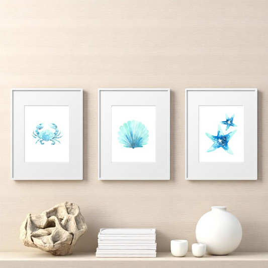 Set of three framed beach-themed art prints in a modern coastal setting, styled above a light wood shelf with stacked books, coral, and minimalist ceramics.