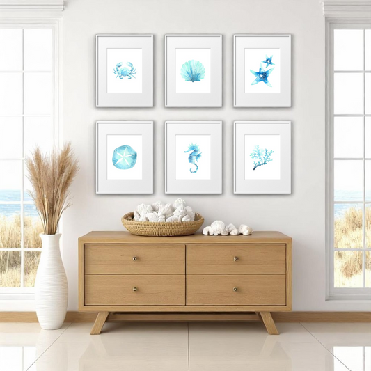 Set of six 8x10 beach-themed art prints in white frames arranged in a 3x2 grid above a natural wood console, styled with a coastal basket of shells, pampas grass, and ocean view windows.