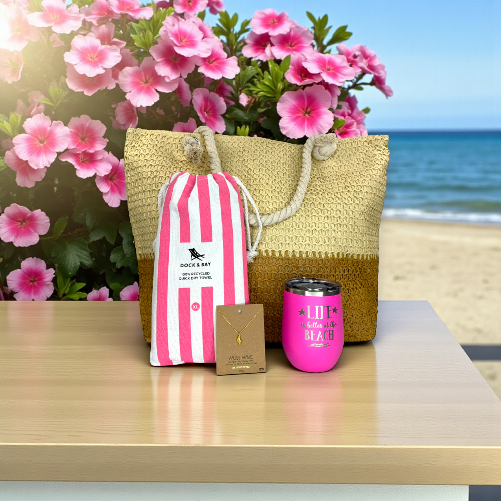 beach lover gift set with pink striped beach towel, pink tumbler, dainty conch shell necklace and straw beach bag on a light wood table with pink tropical flowers and ocean in the background.