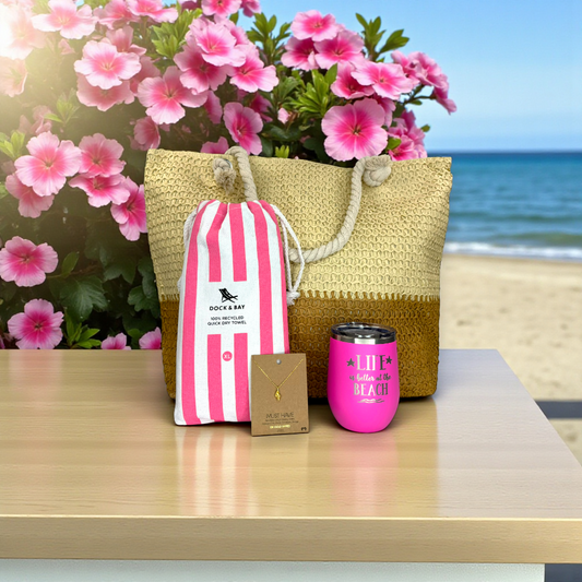beach lover gift set with pink striped beach towel, pink tumbler, dainty conch shell necklace and straw beach bag on a light wood table with pink tropical flowers and ocean in the background.