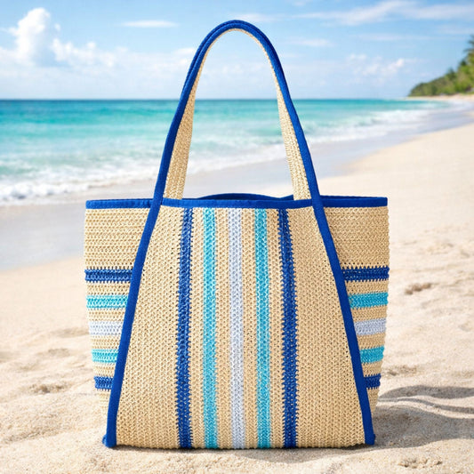Blue and aqua striped straw beach tote bag on sand with ocean background, blue trim and woven straw texture