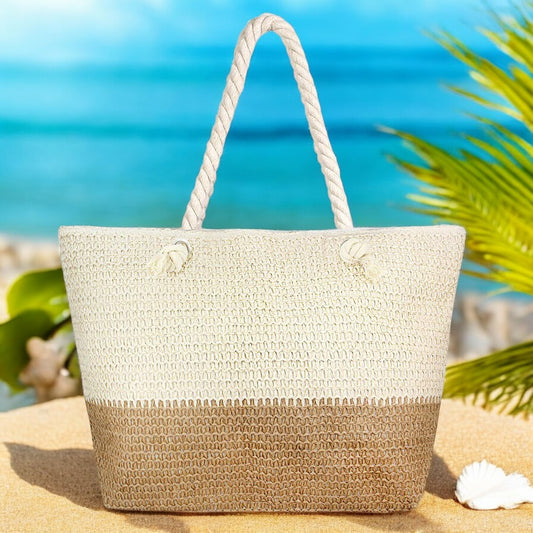 Oversized straw beach tote bag with beige and tan two-tone design on a sandy beach with turquoise ocean in the background.