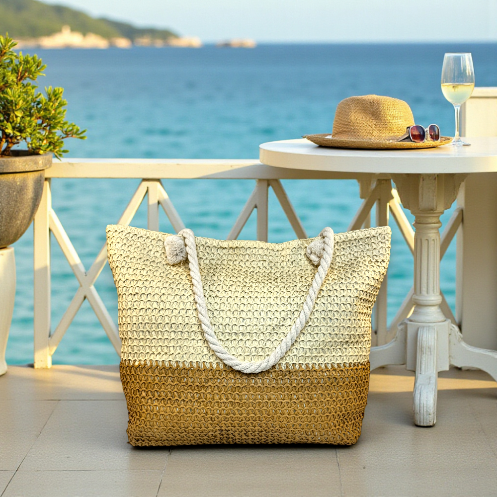 Straw beach bag for women beside a seaside café table with hat and wine glass overlooking the ocean.