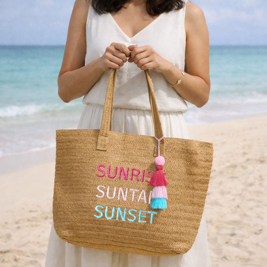 Woman holding Raffia Message Beach Bag Tote: Sunrise, Suntan, Sunset with Tassel standing on a sandy beach with ocean in background.