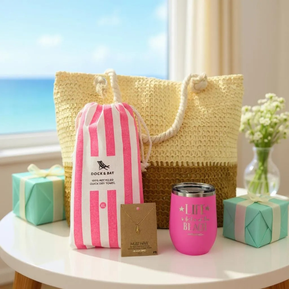 gift table with a beach lover gift set including a straw beach bag, pink tumbler, beach towel gift box, and necklace in a bright room with ocean view.