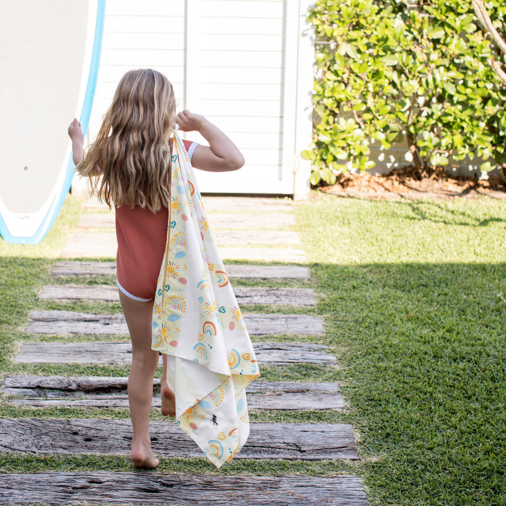 girl walking on wood pathway holding pastel, yellow striped rainbow pattern quick dry sand free kids beach towel
