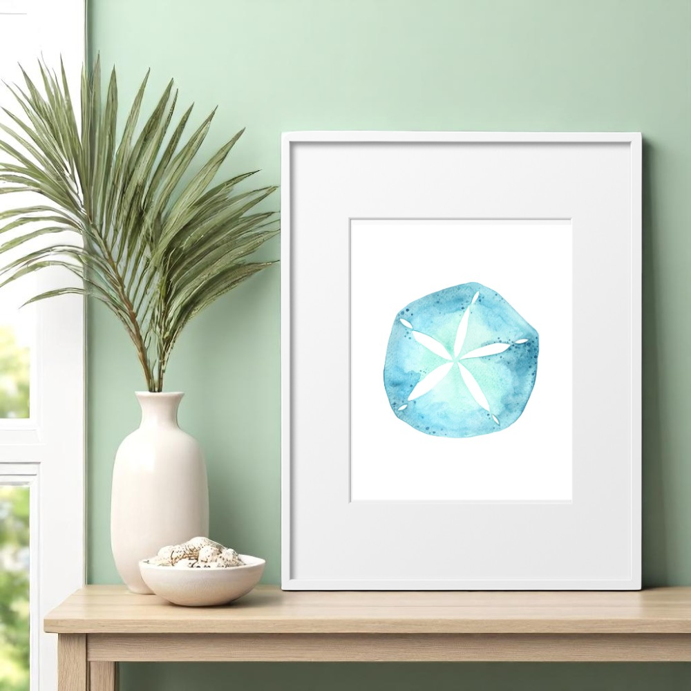 Single 8x10 framed sand dollar art print styled above a light wood console table with a white bowl of seashells and a tall vase of dried palm leaves on the side, creating a calm coastal entryway scene with natural light.