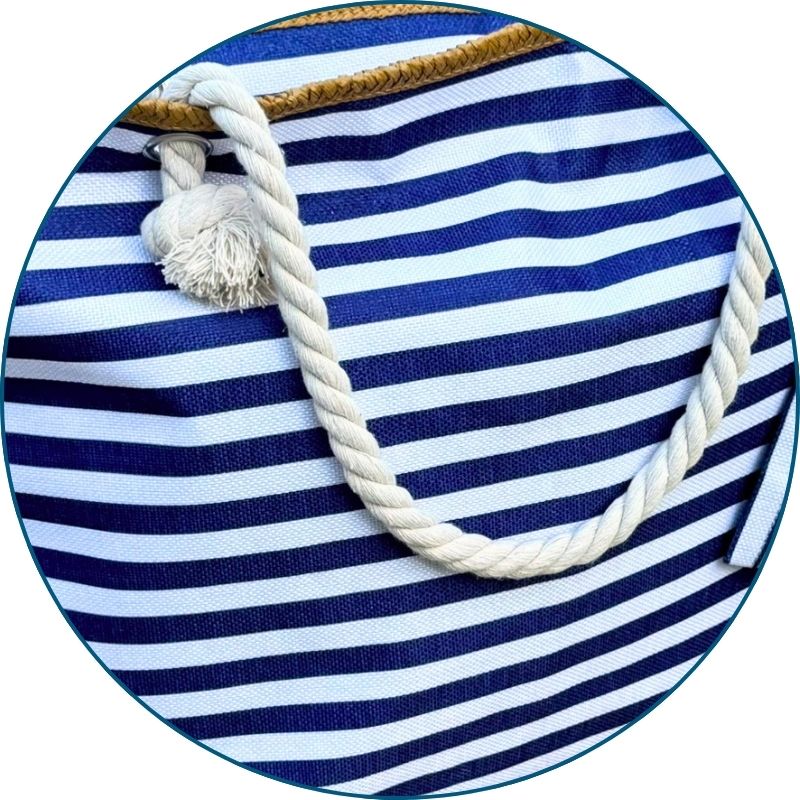 Close-up of soft rope handles on a navy and white striped beach bag against a coastal background.