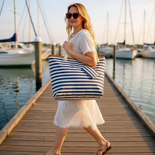 Woman holding a nautical navy and white striped beach bag on a dock with boats in the background