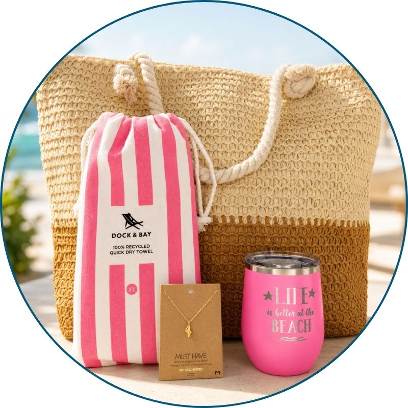Pink beach lover gift set styled by the ocean with straw beach bag, striped towel, pink tumbler and gold-dipped conch shell necklace.