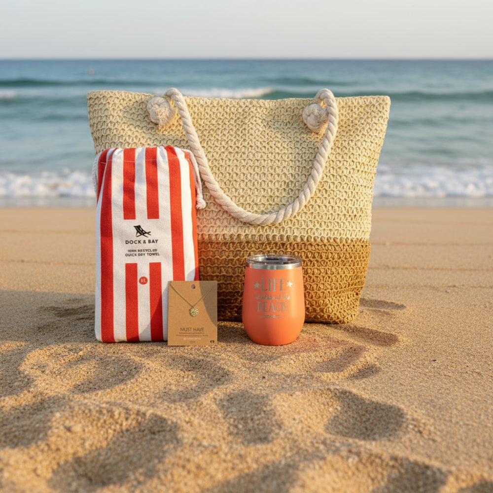 Woven straw bag with burnt orange striped towel, tropical monstera necklace, and peach tumbler on a sandy beach.