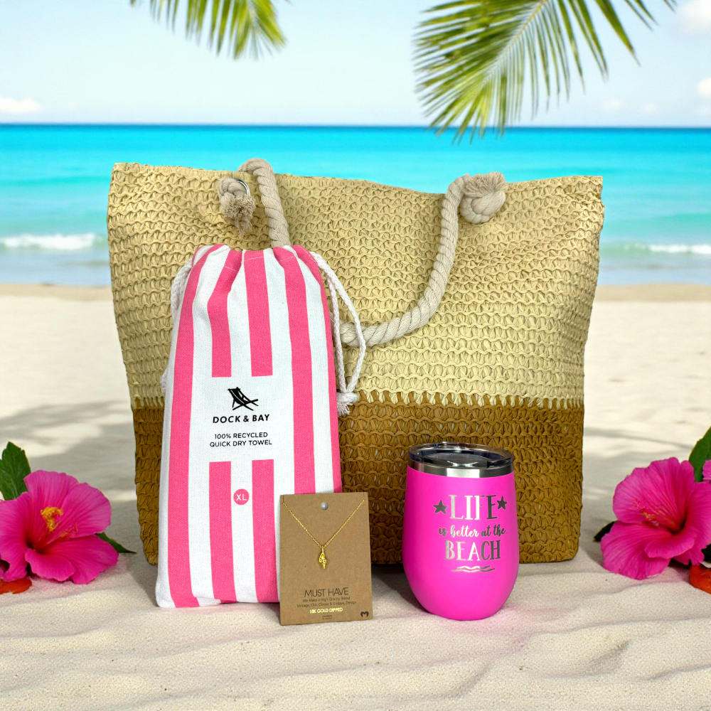 Bright pink beach lover gift set displayed on white sand with turquoise ocean in the background, surrounded by green palm leaves and fuchsia tropical flowers in bright natural sunlight.