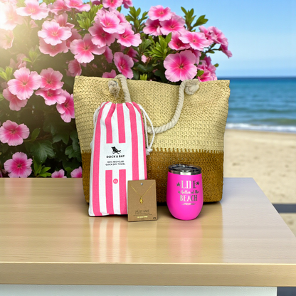 beach lover gift set with pink striped beach towel, pink tumbler, dainty conch shell necklace and straw beach bag on a light wood table with pink tropical flowers and ocean in the background.