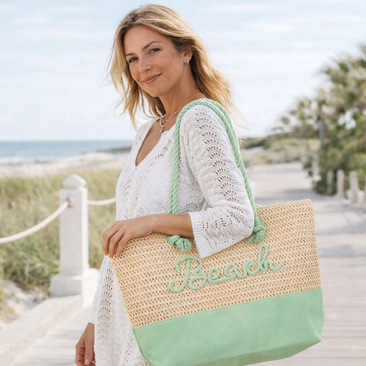 Woman holding mint green straw beach tote bag with raised “Beach” lettering on a boardwalk at the beach