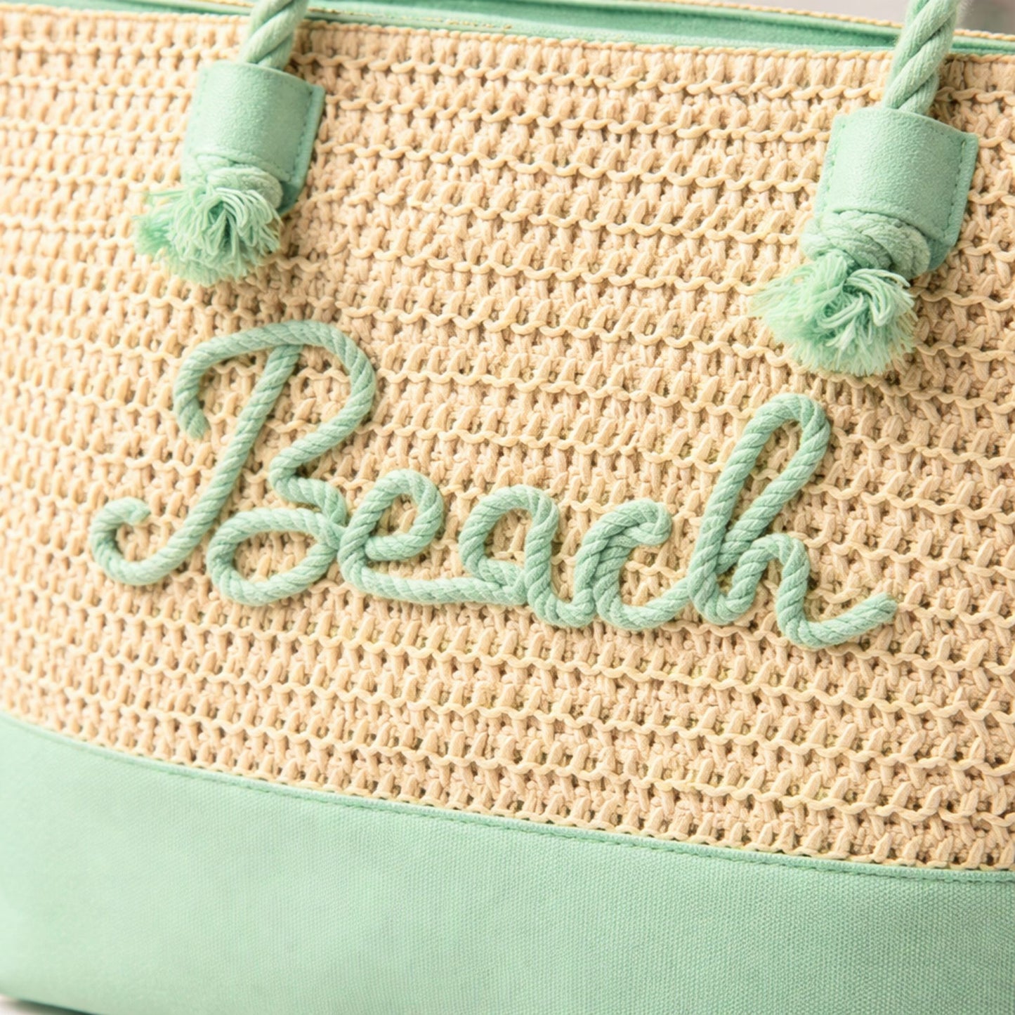 Close-up of mint green raised “Beach” lettering, rope handles, and zipper on straw tote bag