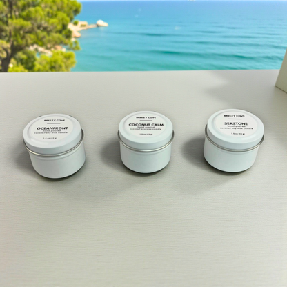 Three Breezy Cove 1.5 oz coconut soy wax mini white tin candles—Oceanfront, Coconut Calm, and Seastone—displayed on a light surface with a coastal ocean view in the background.