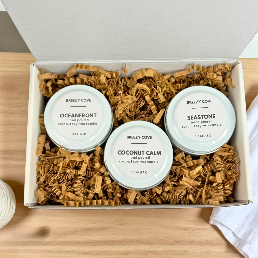 Open white gift box filled with crinkle paper, featuring three Breezy Cove 1.5 oz coconut soy wax mini candles in Oceanfront, Coconut Calm, and Seastone scents arranged neatly inside.