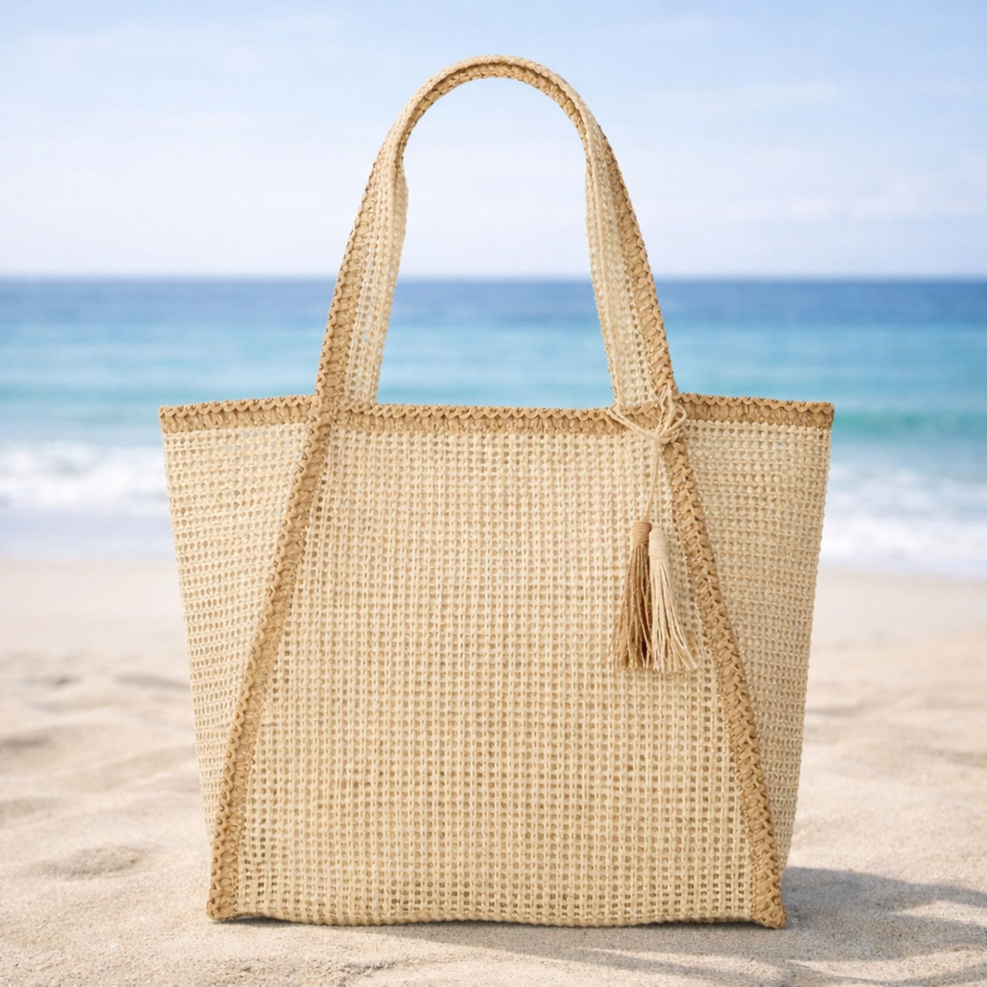 Beige woven straw beach bag tote with magnetic closure on sand with ocean in the background.
