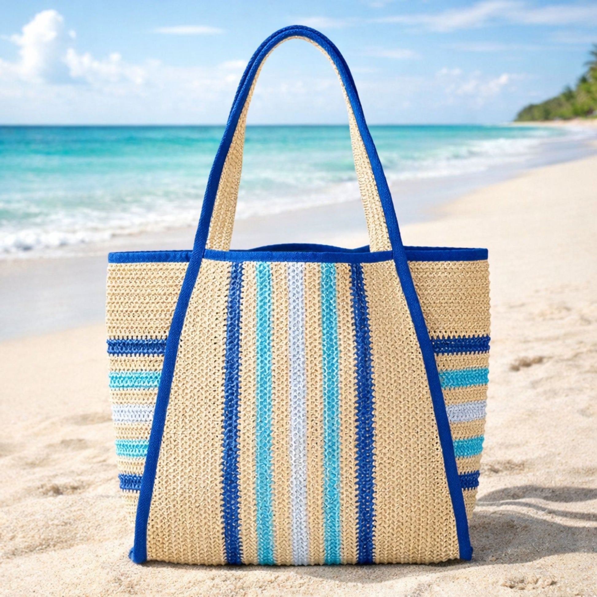 Blue and aqua striped straw beach tote bag on sand with ocean background, blue trim and woven straw texture