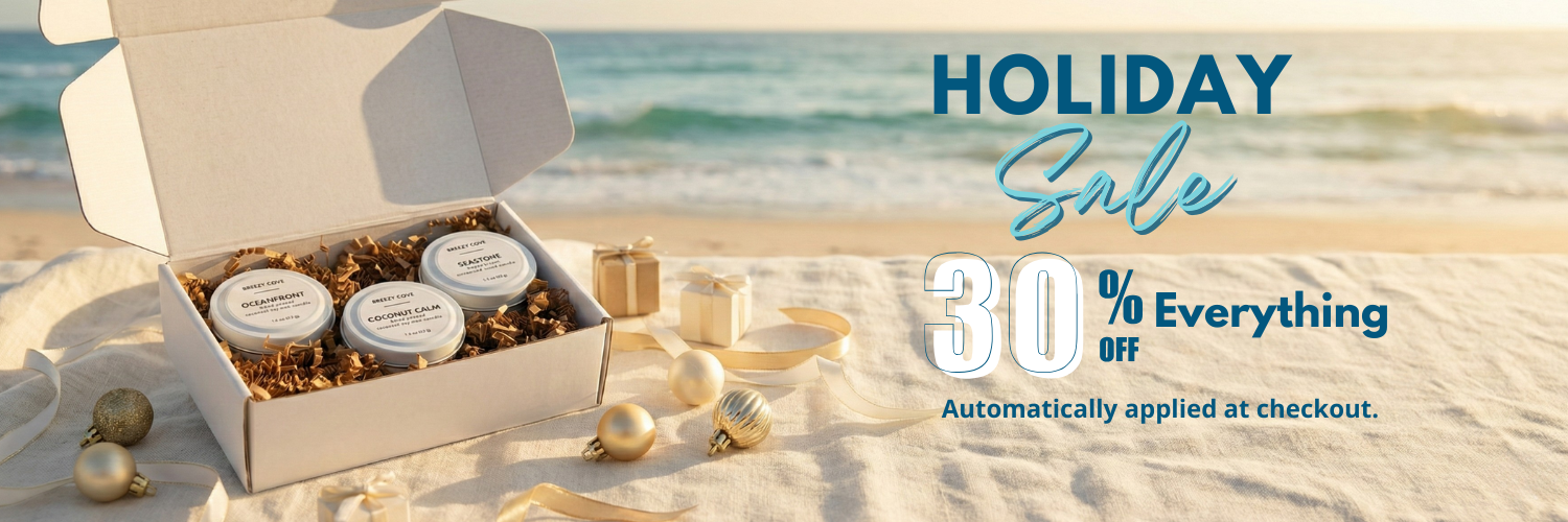 Beachside mini candle trio gift set in an open box resting on an ivory linen blanket with gold and ivory ornaments and small gift boxes, ocean waves in the background — a cozy coastal Christmas scene featuring gifts for beach lovers and the perfect beach lover gift idea.