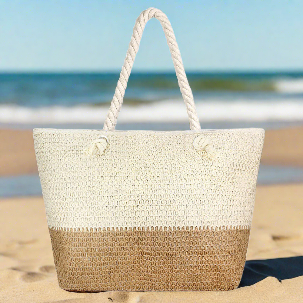 large, oversize beige and tan straw beach bag with zipper and rope handles on sandy beach with ocean