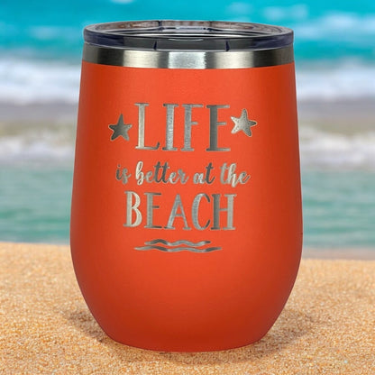 Coral "Life is Better at the Beach" tumbler resting on the sand, perfect for beach lovers and keeping drinks cool or warm