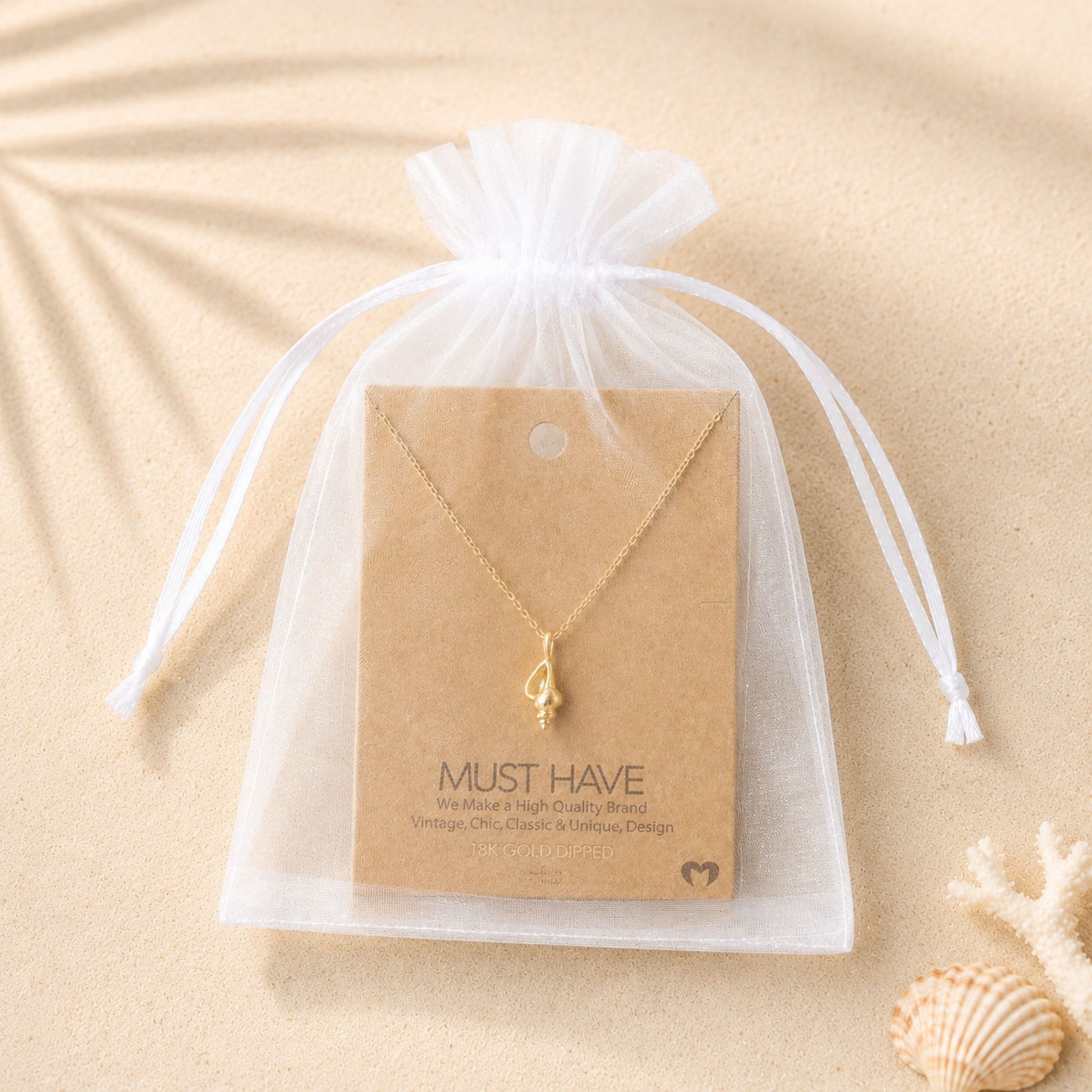 18k gold dipped conch shell necklace displayed inside a white organza gift bag on a soft sandy beach-inspired background.