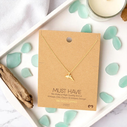 Dolphin gold pendant necklace on a jewelry card styled on a vanity tray with sea glass, driftwood, and a candle, set against a light coastal background in natural sunlight.