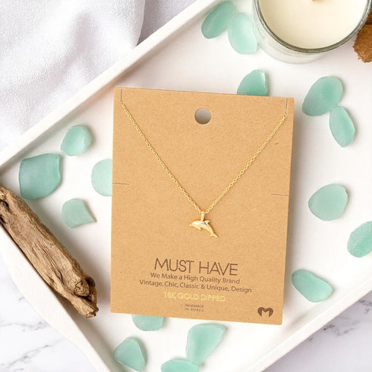 Dolphin gold pendant necklace on a jewelry card styled on a vanity tray with sea glass, driftwood, and a candle, set against a light coastal background in natural sunlight.