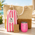 Pink beach lover gift set with straw beach bag, pink striped quick dry towel, pink tumbler, and gold dipped conch shell necklace in a sunny beach resort setting