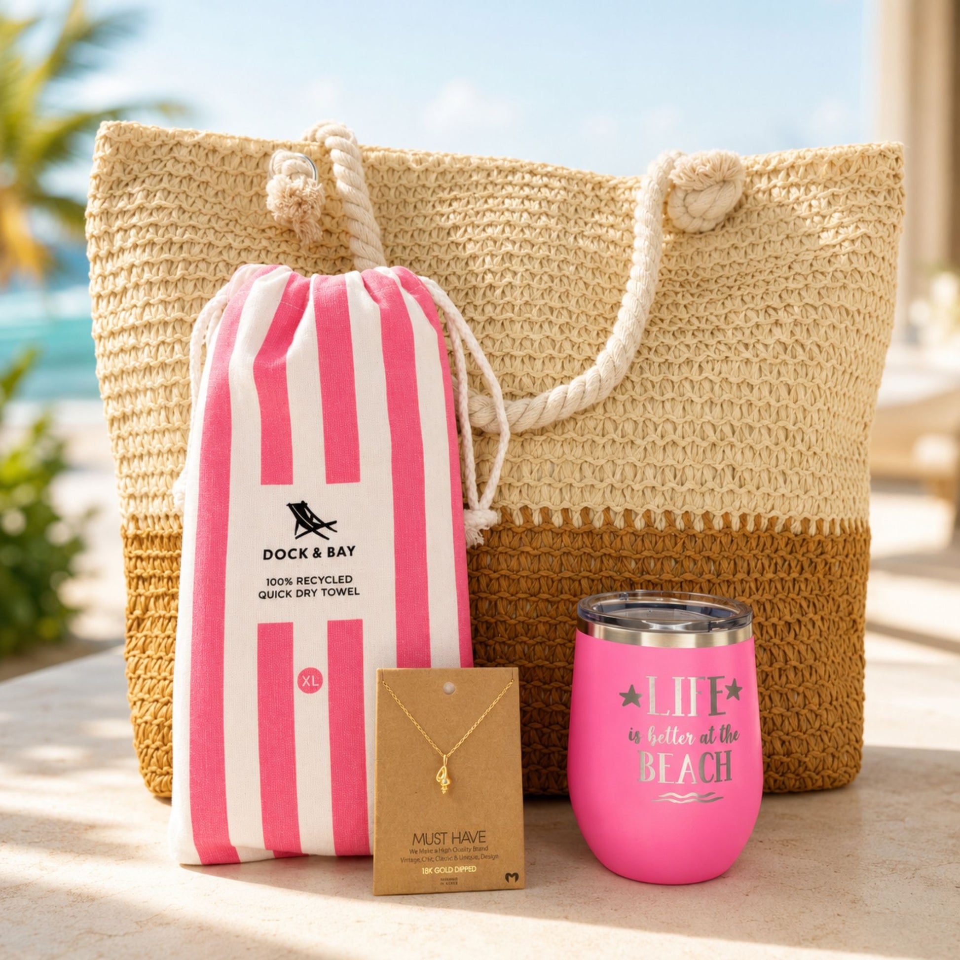 Pink beach lover gift set with straw beach bag, pink striped quick dry towel, pink tumbler, and gold dipped conch shell necklace in a sunny beach resort setting
