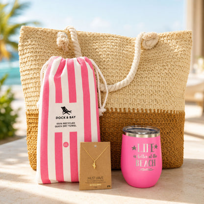 Pink beach lover gift set with straw beach bag, pink striped quick dry towel, pink tumbler, and gold dipped conch shell necklace in a sunny beach resort setting