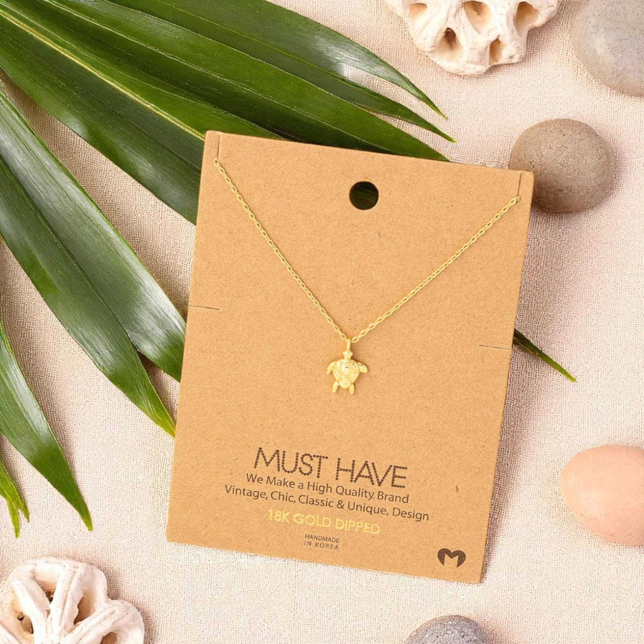 Gold sea turtle pendant necklace on a jewelry card in a flat lay with coral pieces, smooth beach stones, and a palm leaf, styled on natural linen