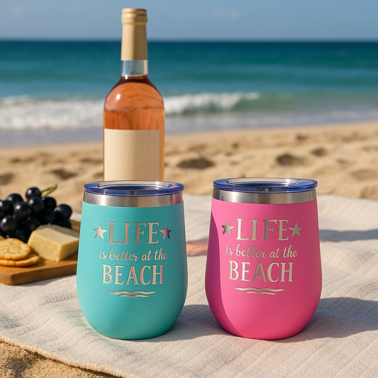 Teal and pink insulated beach tumblers on a picnic blanket with snacks and drinks, sand and sea in the background.