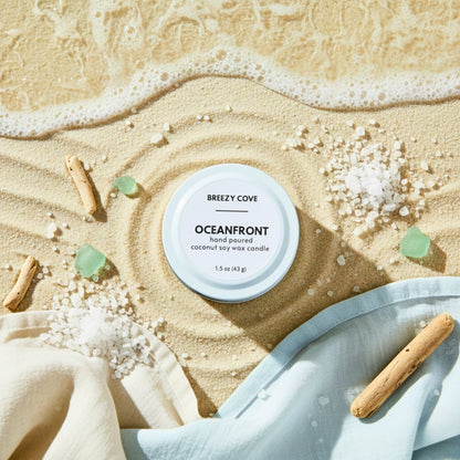 Oceanfront beach scented candle on sand with sea glass and driftwood near the shoreline.