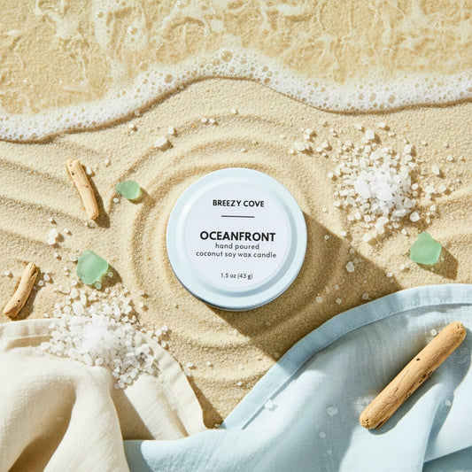 Oceanfront beach scented candle on sand with sea glass and driftwood near the shoreline.
