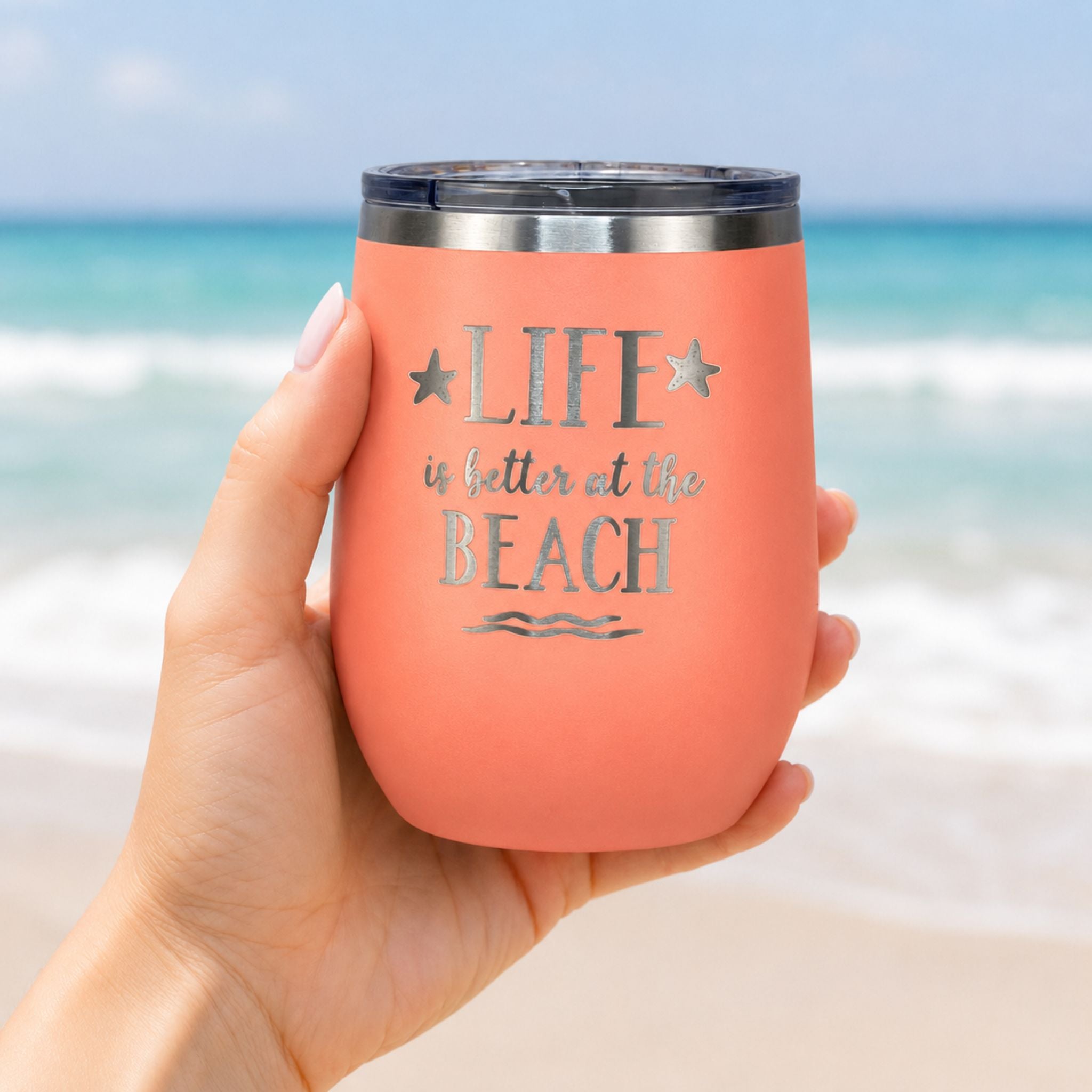 Hand holding peach beach wine tumbler with lid in front of the ocean