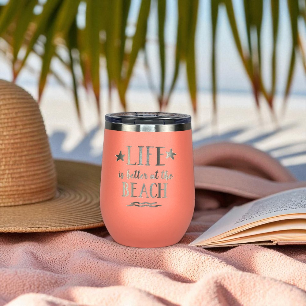 peach metal wine tumbler with 'Life is better at the beach' text on a beach setting with a hat and book.