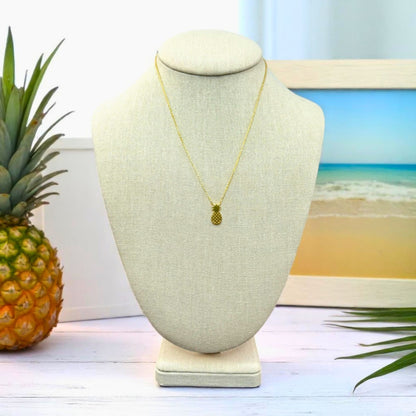 18k gold dipped pineapple pendant necklace displayed on a linen jewelry bust with tropical beach-inspired decor.