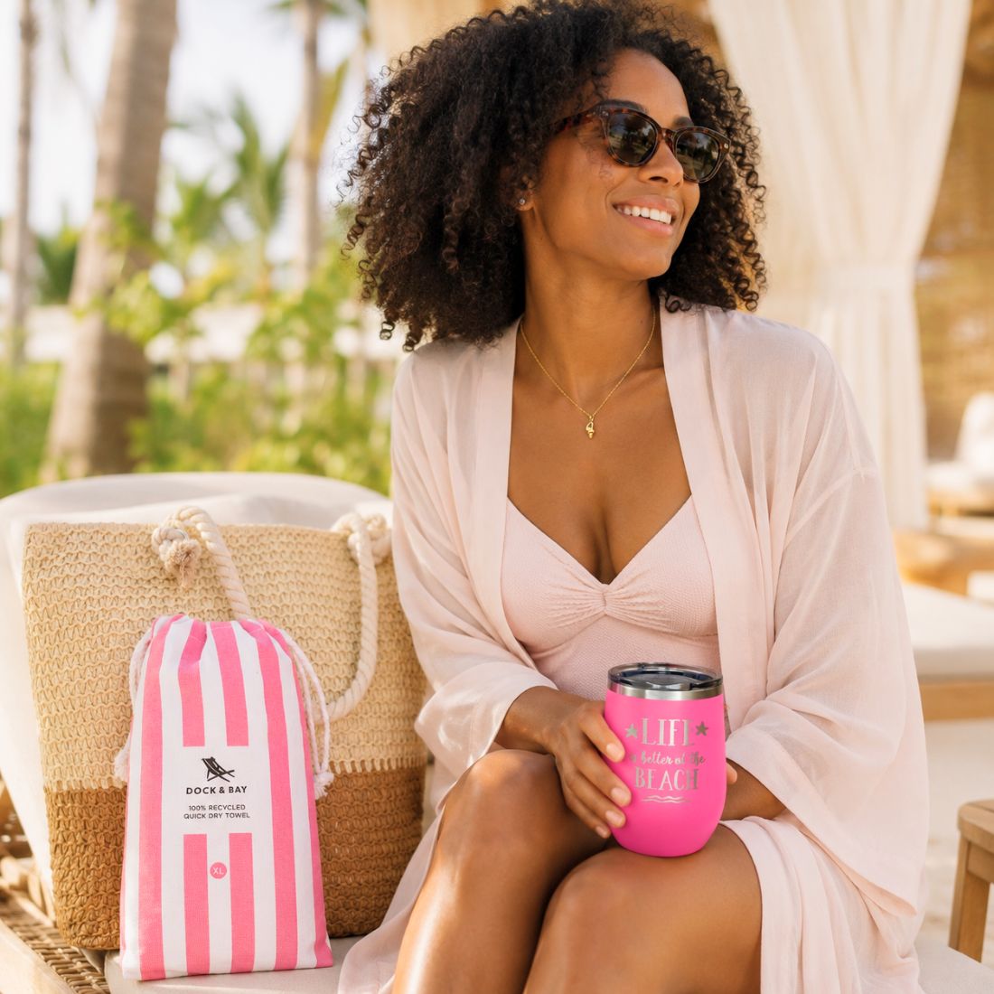 Woman relaxing in a resort cabana with pink beach lover gift set, straw beach bag, striped towel, pink tumbler and gold-dipped conch shell necklace.