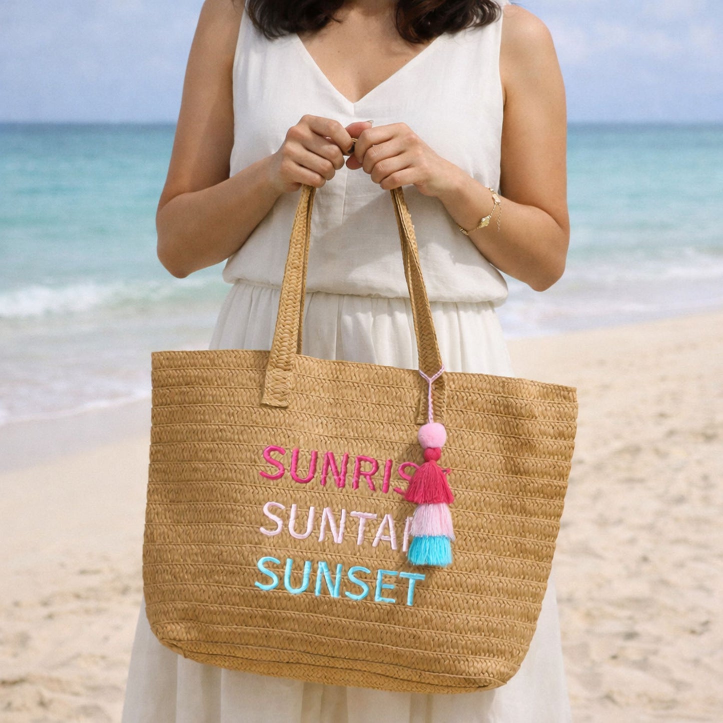 Woman holding Raffia Message Beach Bag Tote: Sunrise, Suntan, Sunset with Tassel standing on a sandy beach with ocean in background.
