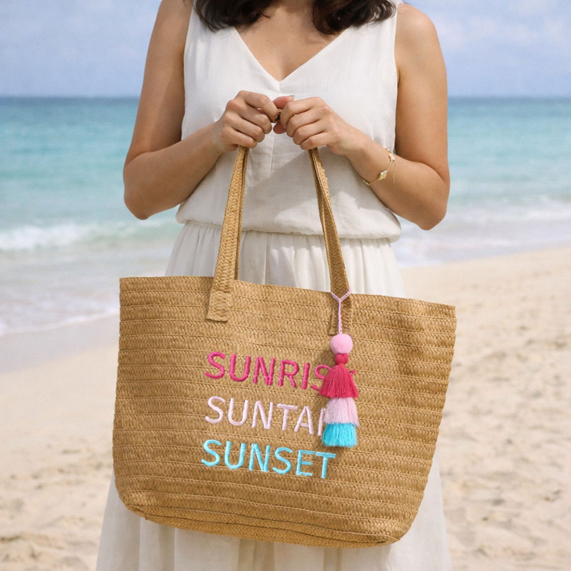 Woman holding Raffia Message Beach Bag Tote: Sunrise, Suntan, Sunset with Tassel standing on a sandy beach with ocean in background.