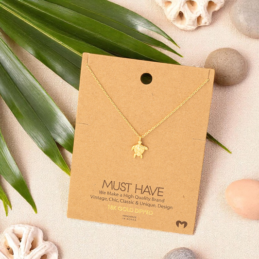 Sea turtle pendant necklace displayed on a jewelry card in a flat lay with coral pieces, smooth beach stones, and a palm leaf, styled on sand-toned linen with soft natural light.