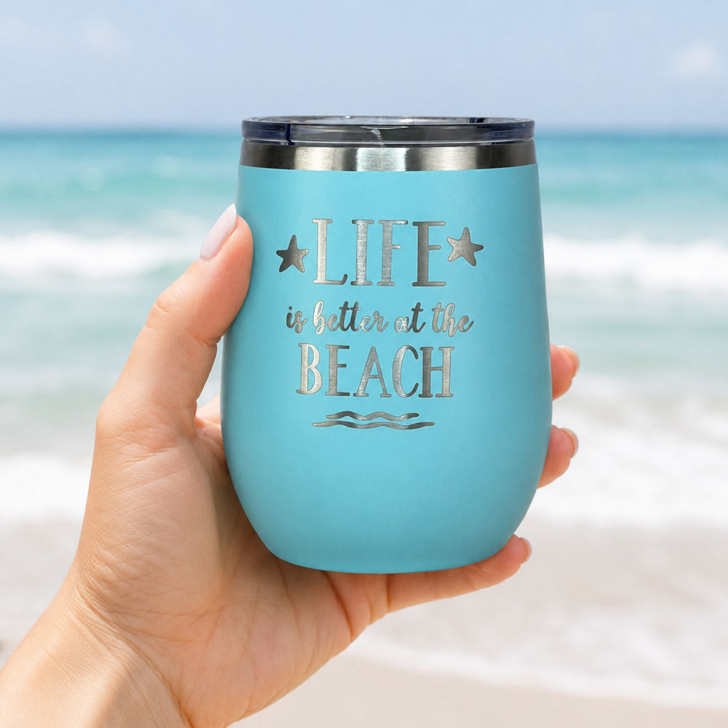 Hand holding teal beach wine tumbler with lid in front of an ocean beach background
